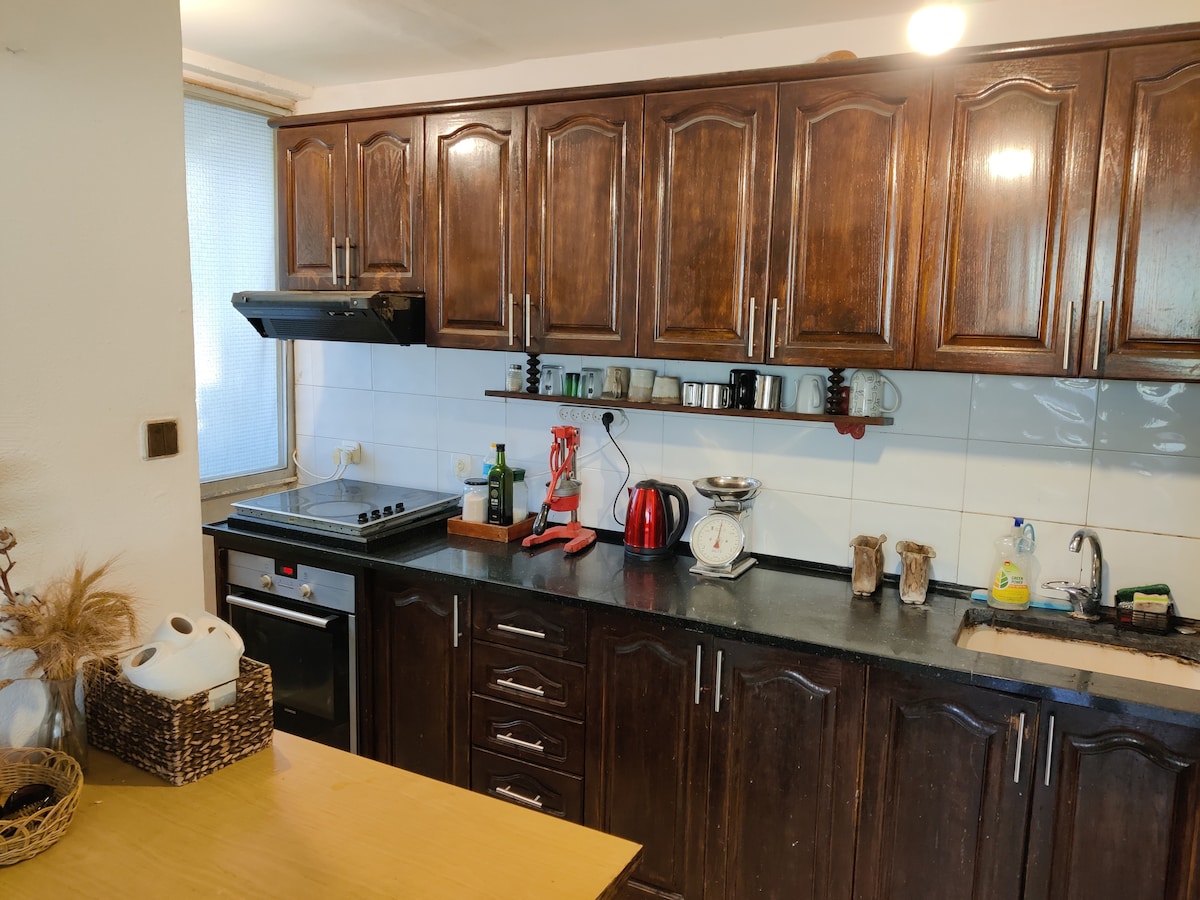 The kitchen features dark wooden cabinetry with a spacious countertop. An array of kitchen utensils, a kettle, and a coffee maker are neatly arranged. A window allows natural light to enter, illuminating the cooking space. Appliances include a stove and an oven.