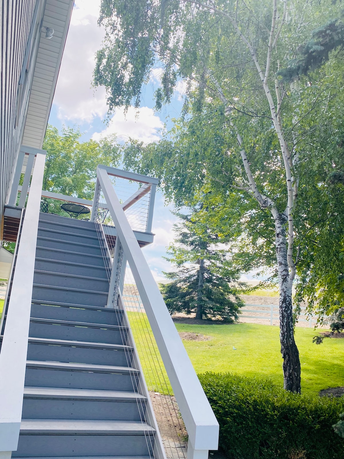 A set of outdoor stairs leads to a separate entrance, flanked by lush greenery. The structure is surrounded by well-maintained lawns and tall trees, with a clear blue sky and soft clouds in the background, offering a peaceful, natural setting.