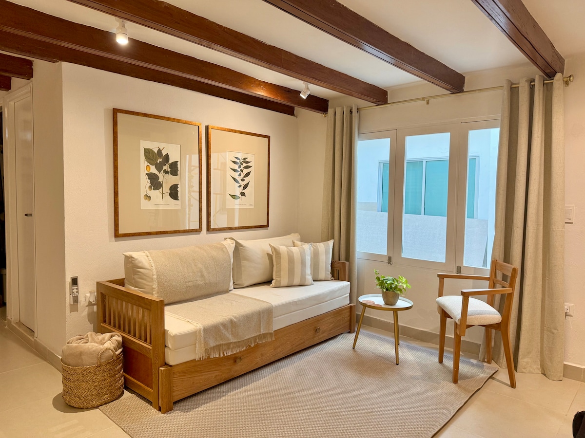A light-filled living area features a wooden sofa adorned with neutral-toned cushions, accompanied by a sleek armchair. Two botanical prints hang above the couch, while potted greenery sits on a small table. Large windows invite natural light, complementing the warm wood beams overhead.