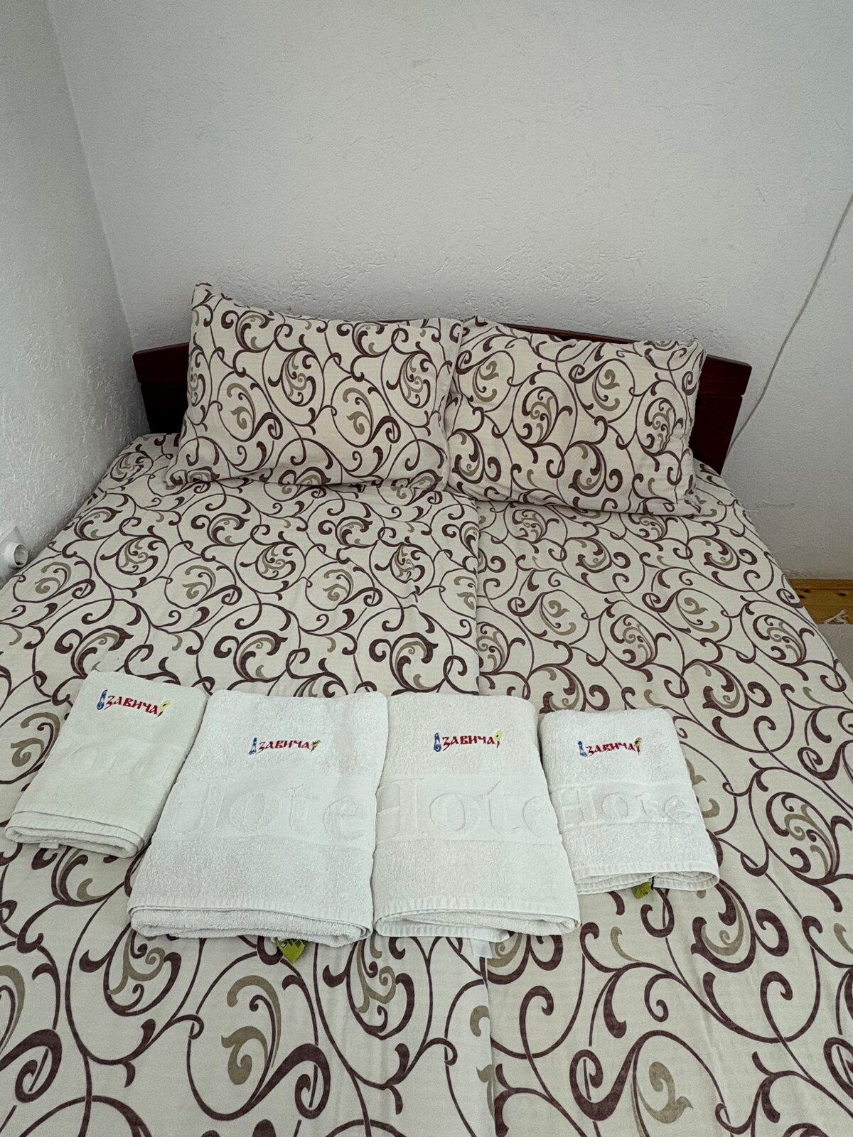 The sleeping area features a double bed with an intricate patterned bedspread and two matching decorative pillows. Four neatly arranged towels are positioned at the foot of the bed, displaying a logo. The surrounding walls are in a simple light color.
