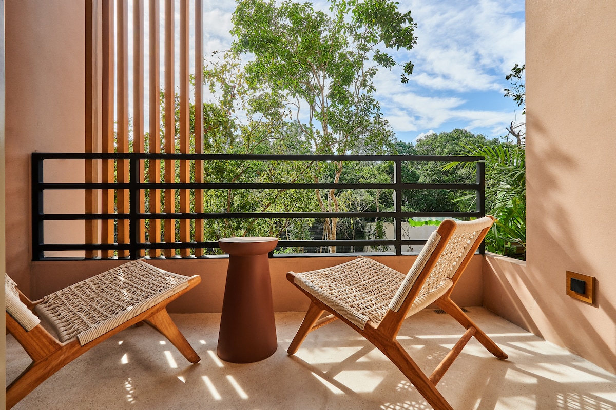 A private terrace features two wooden lounge chairs and a round side table. Lush greenery is visible through the railing, creating a peaceful setting. Natural light brightens the space, highlighting the earthy tones of the terrace walls.
