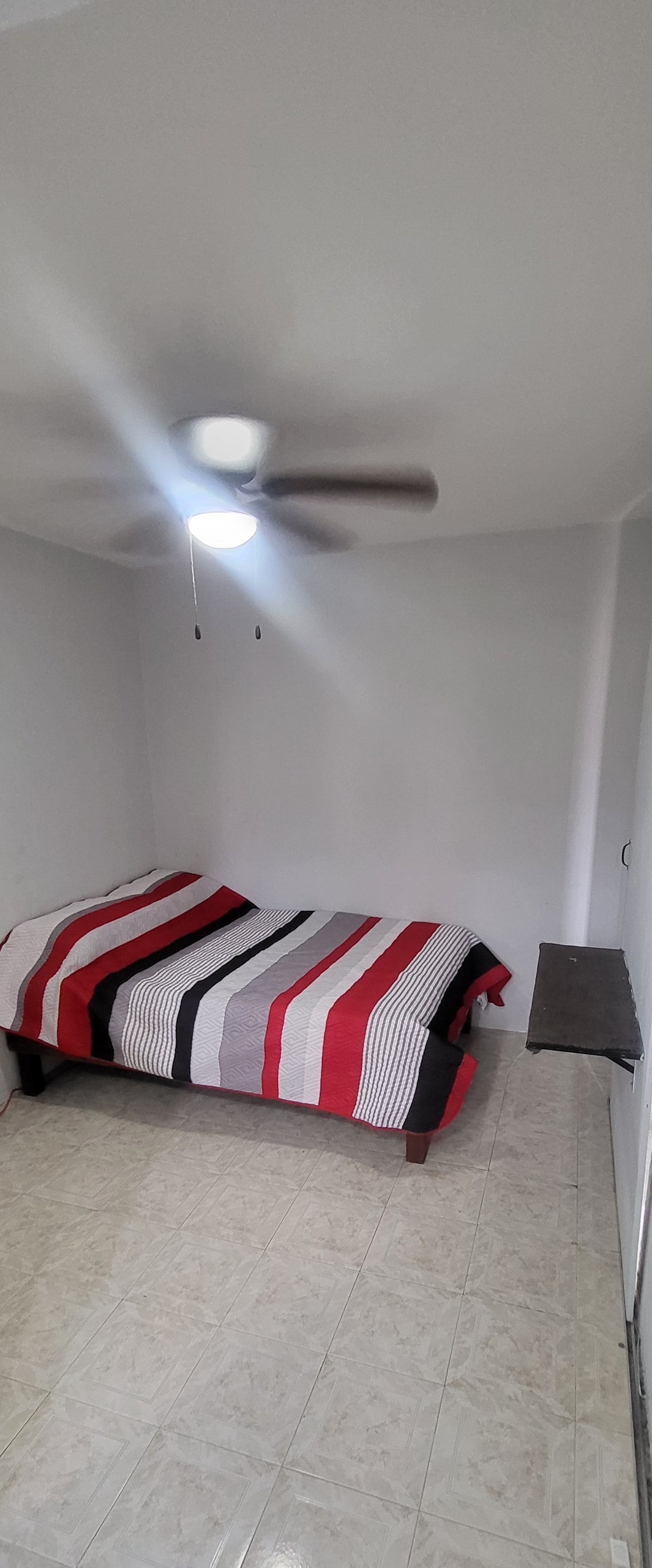 A spacious sleeping area features a double bed dressed in a striped red and black comforter. A fan hangs from the ceiling, while a small table is positioned along one wall. Light-colored tiles cover the floor, contributing to a calm environment.