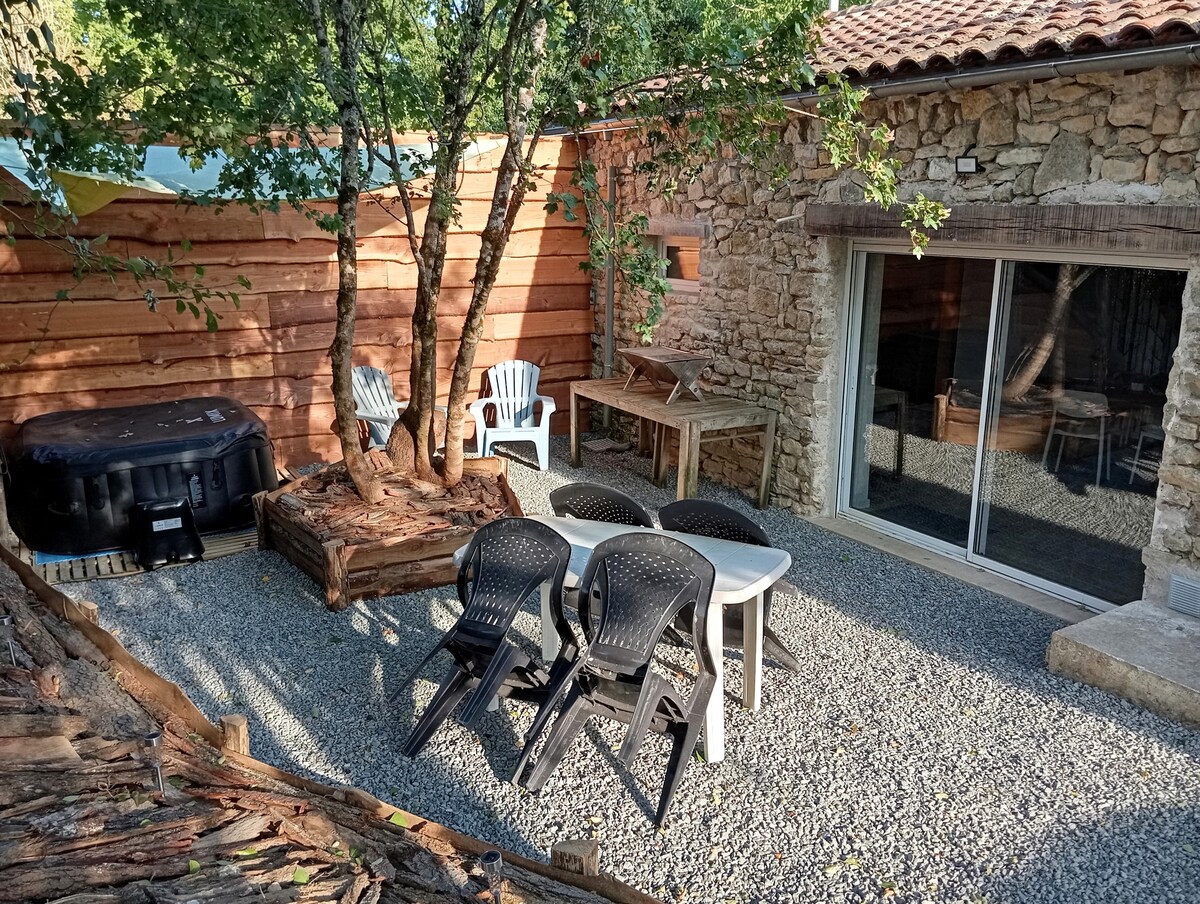 An outdoor area features a gravel surface surrounded by natural elements. A wooden table and six black plastic chairs are set under the shade of trees. A hot tub and additional seating options are visible, with rustic stone walls providing a charming backdrop.