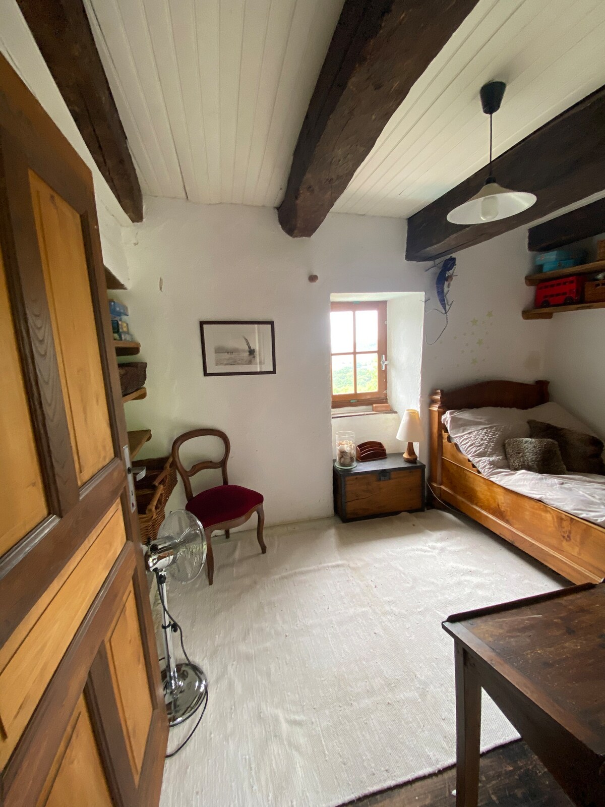 A simple and charming room features exposed wooden beams and a light-painted ceiling. A single bed with a textured blanket is positioned against the wall, beside a window that allows natural light to illuminate the space. A small desk and a red chair add functional elements.