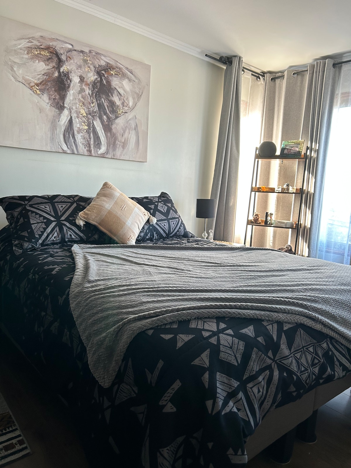 A cozy bedroom features a spacious bed adorned with a patterned black and white bedspread. An accent pillow is placed on the bed, complemented by a decorative gray throw. Natural light filters through sheer curtains framing a window, and a small bookshelf displays various decorative items.