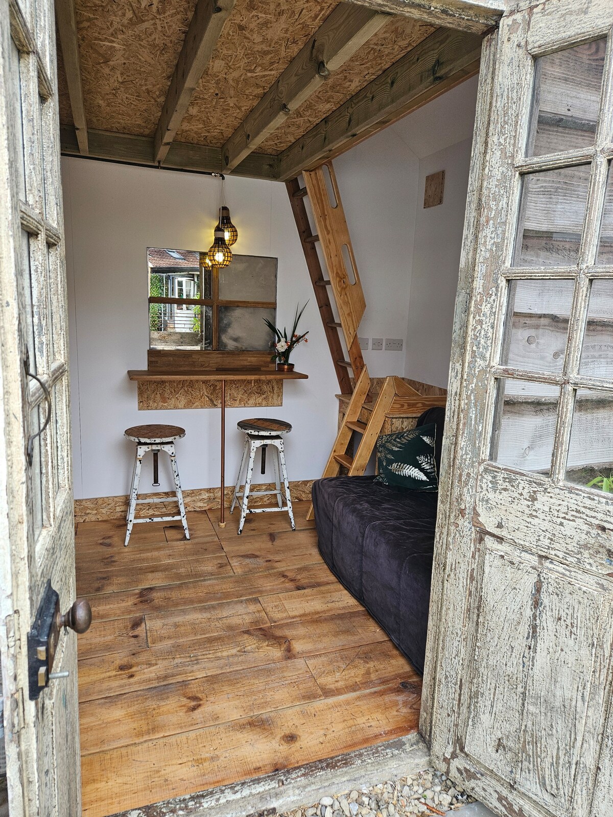 The entrance way reveals a cozy interior of The Den, featuring a timber floor and a compact seating area with a dark sofa. A wooden bar counter with two stools is positioned against the wall, while a ladder leads to the sleeping area above.