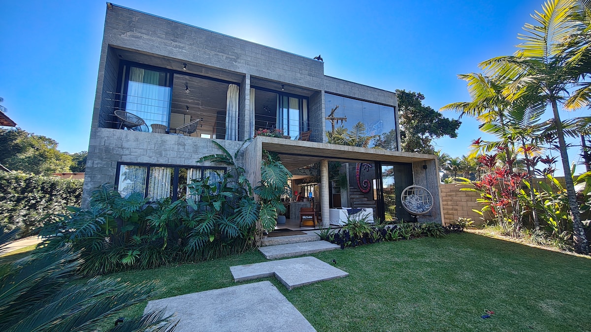 A contemporary two-story house is surrounded by lush greenery. Large glass windows allow natural light to pour into the space. A variety of tropical plants frame the entryway, while a stone pathway leads to the front door. The structure integrates with the natural landscape.