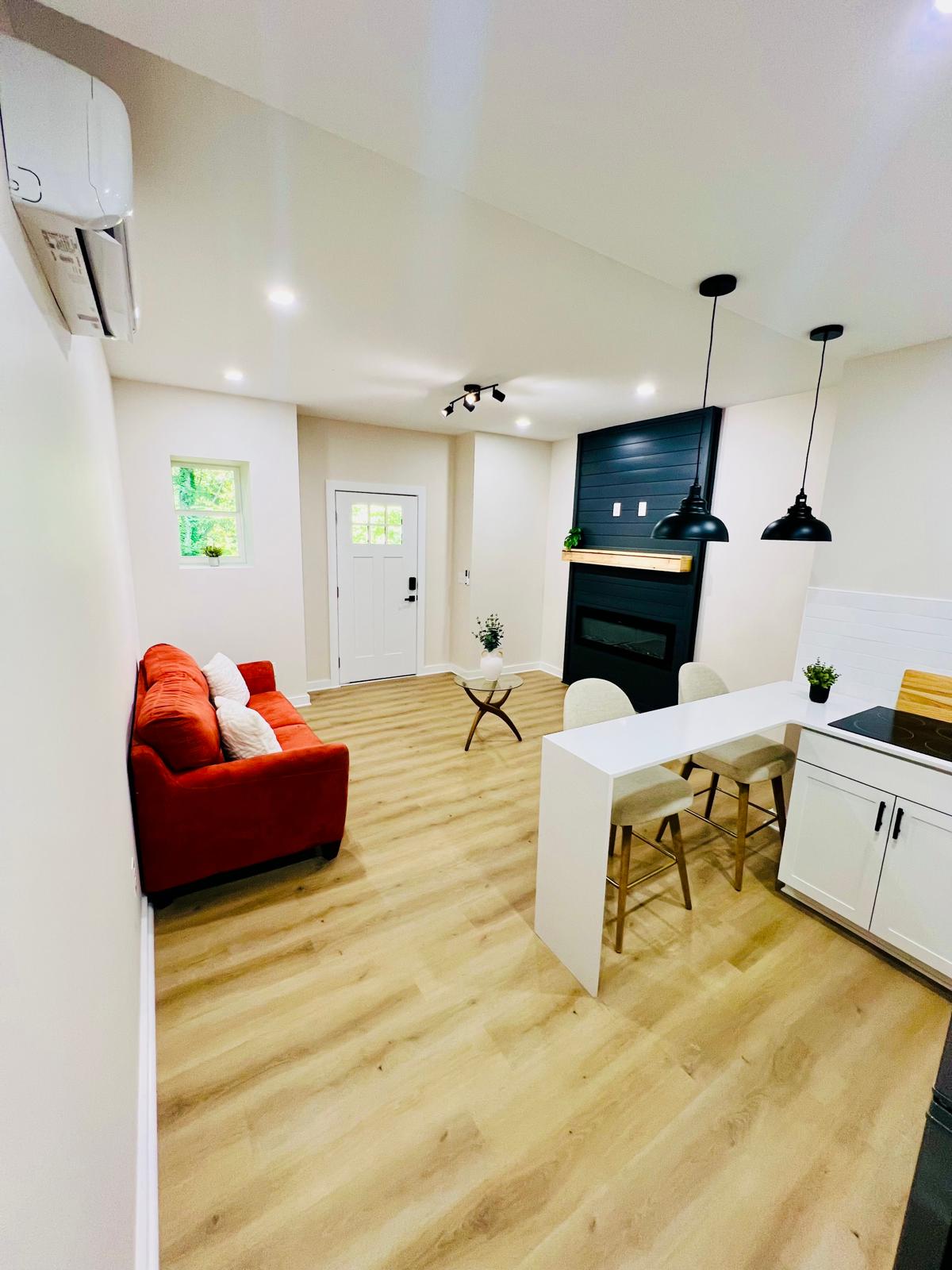 A bright living space features a red sofa with decorative pillows, a small round table, and sleek bar stools at the kitchen counter. A modern fireplace is positioned against a dark wall, while light streams through a nearby window, enhancing the open layout.