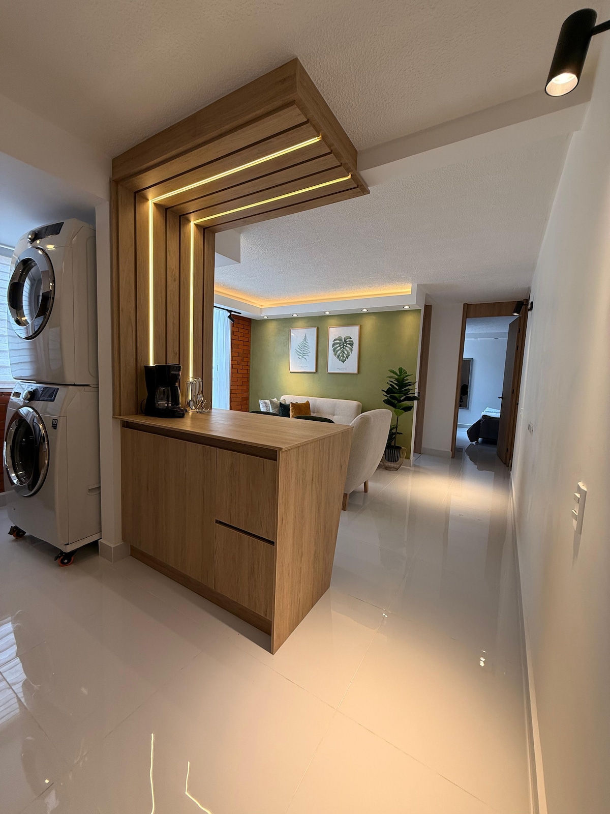 A sleek kitchen area is highlighted by warm wooden accents against a light-colored wall. Modern appliances are visible, including a washer and dryer, while a minimalist coffee station is arranged on the counter. Natural light brightens the airy space, enhancing the open layout.