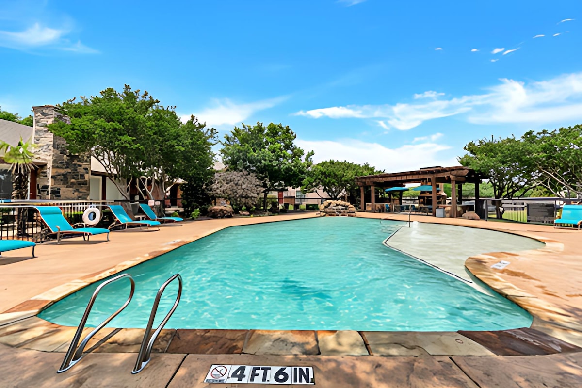 A refreshing swimming pool is framed by lounge chairs and trees, providing a serene outdoor space. A covered pavilion is visible in the background, adding to the communal atmosphere. The pool's shallow area is marked with a depth sign for safety.