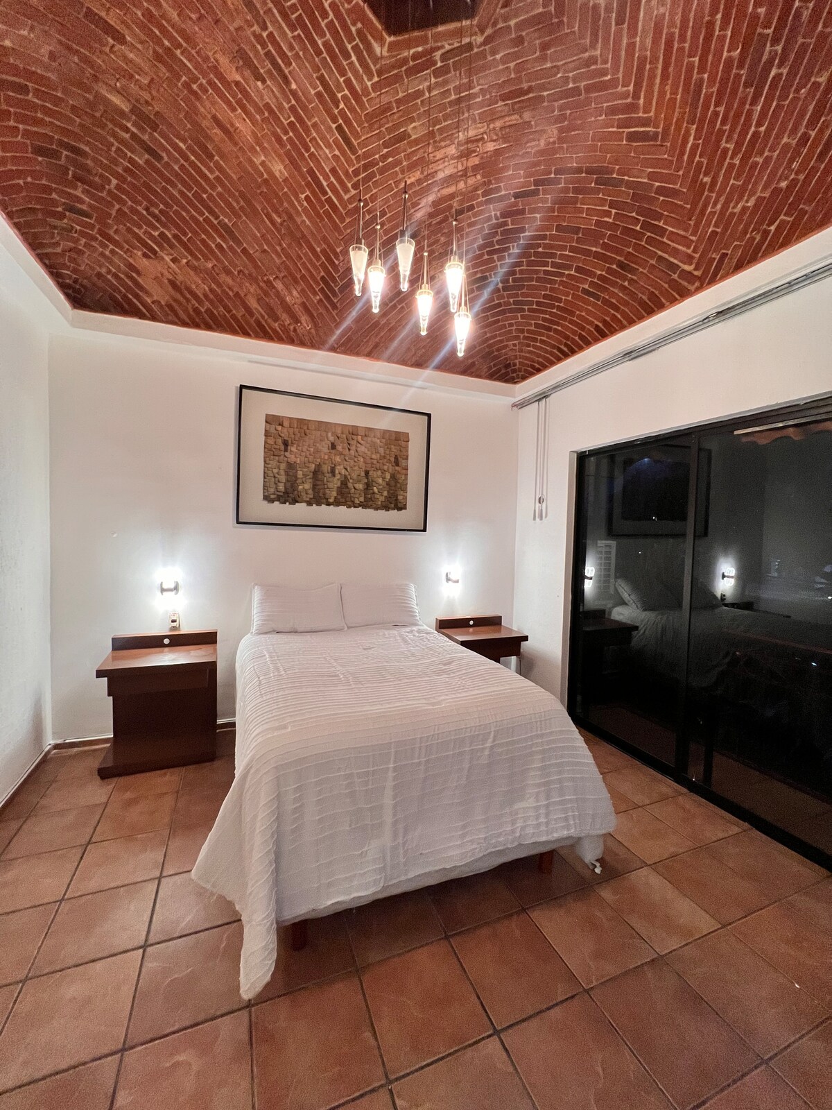 A serene bedroom features a spacious bed with a white blanket, flanked by wooden nightstands. The ceiling is adorned with an intricate brick design, complemented by soft lighting from a modern fixture. Large glass doors offer a view of the outside while allowing natural light.