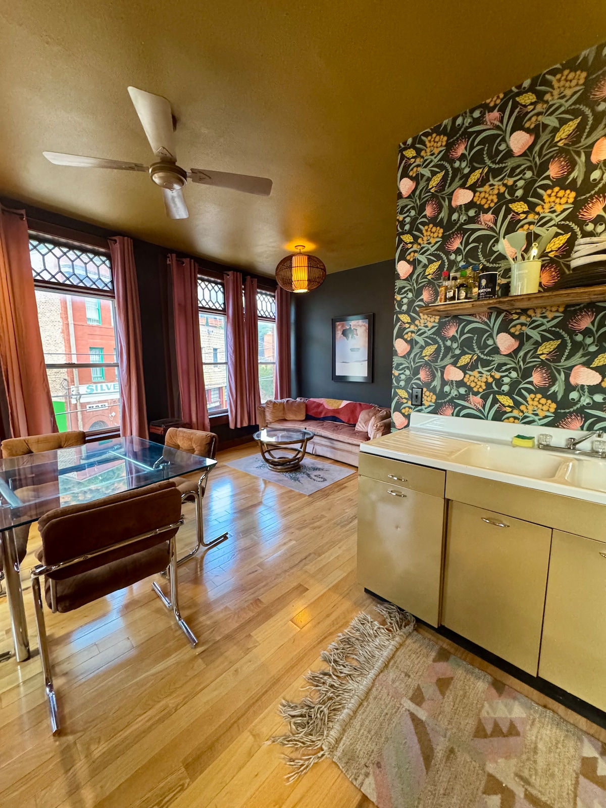 An open living area features large windows adorned with decorative grilles, allowing natural light to fill the space. A cozy seating arrangement with a colorful couch complements the rich wallpaper. A dining table and chairs are situated nearby, while a small kitchen area is visible.
