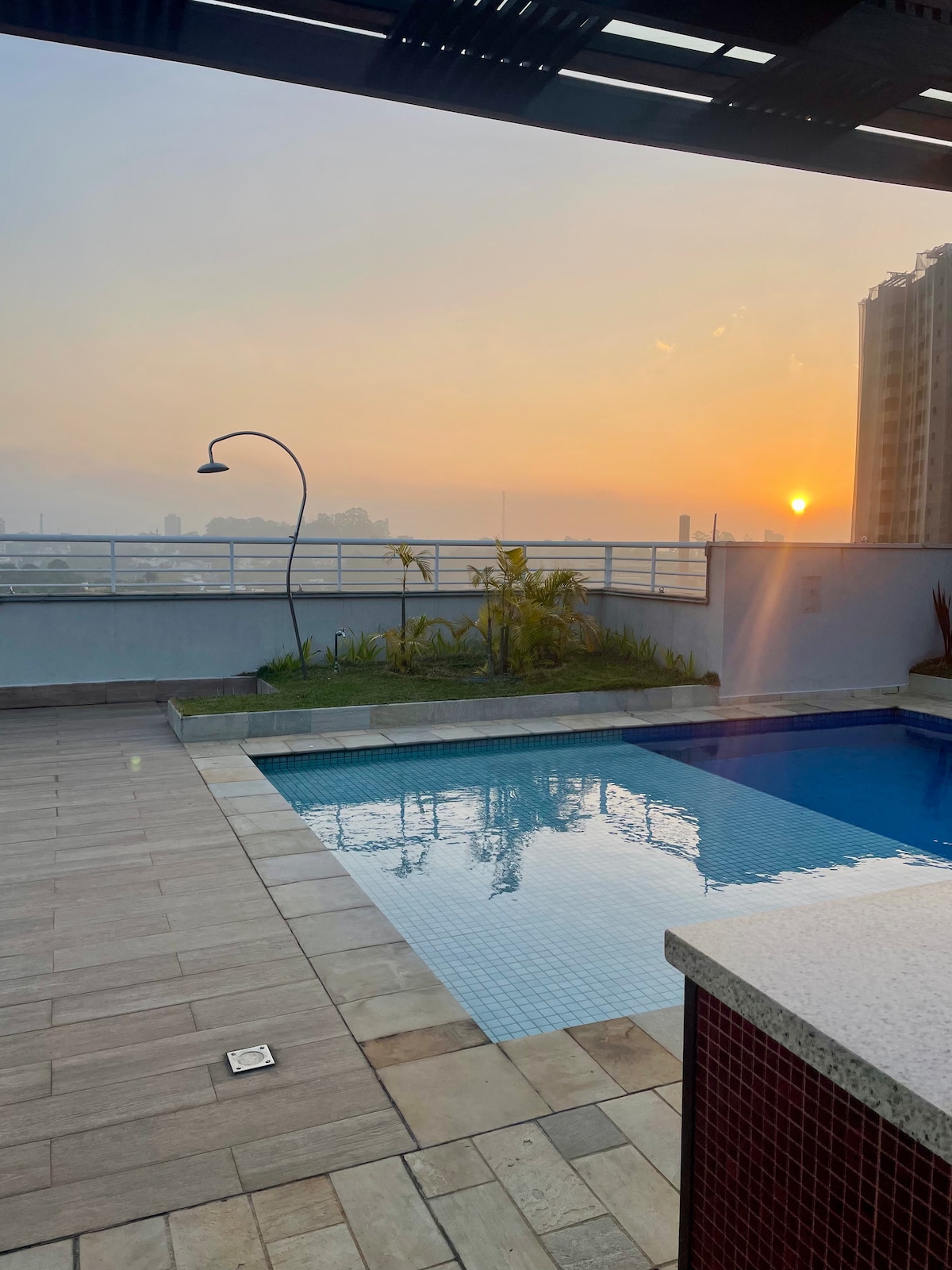 A rooftop pool area is visible at sunset, with the still water reflecting warm hues of orange and yellow. Surrounding greenery adds a natural touch, while the modern building provides a backdrop. The space features a clean deck and a freestanding shower near the pool.