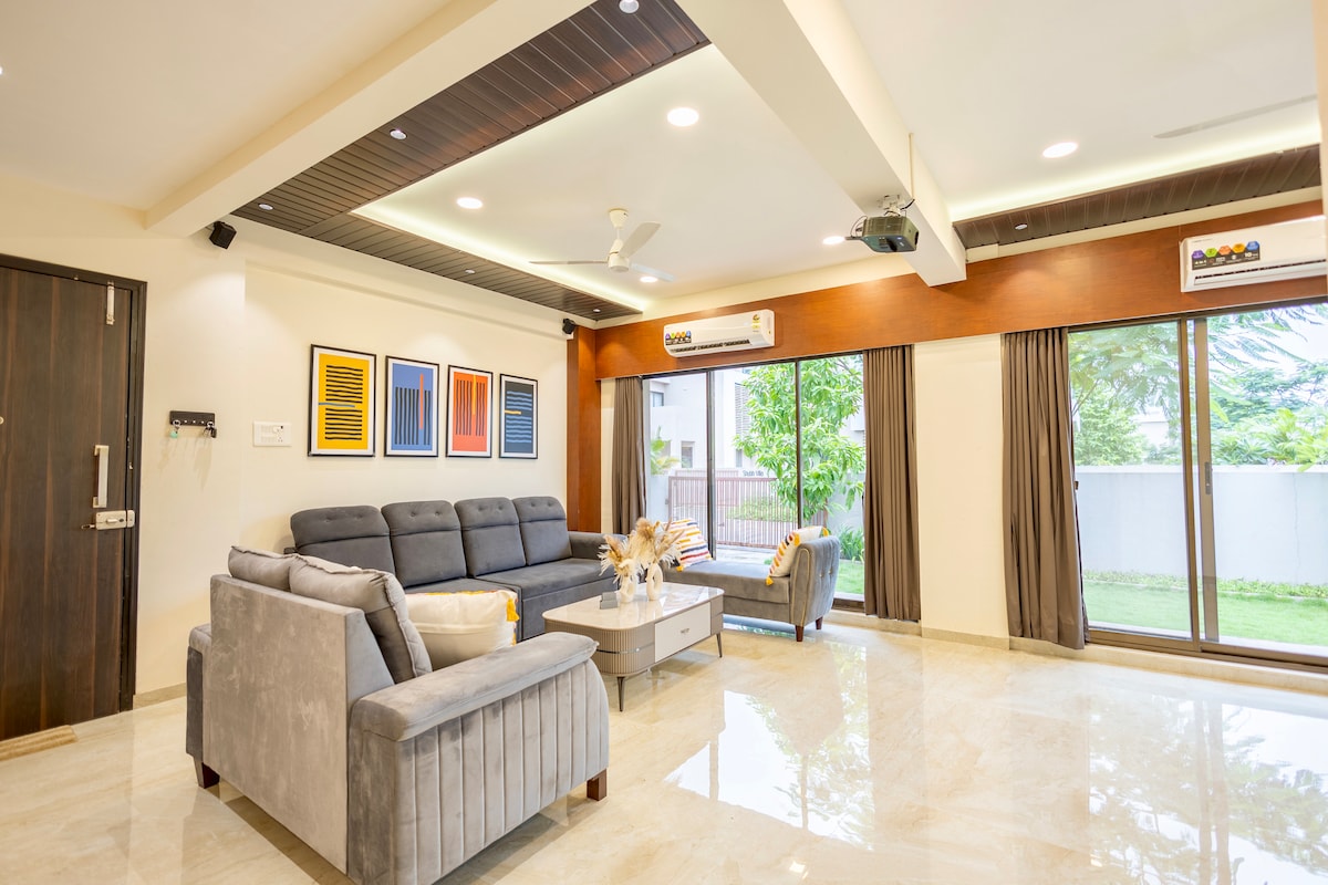 The living area features a spacious setup with comfortable gray sofas and a low coffee table. Large glass doors invite natural light and provide a view of the greenery outside. Wall art adds a pop of color, enhancing the minimalistic decor.