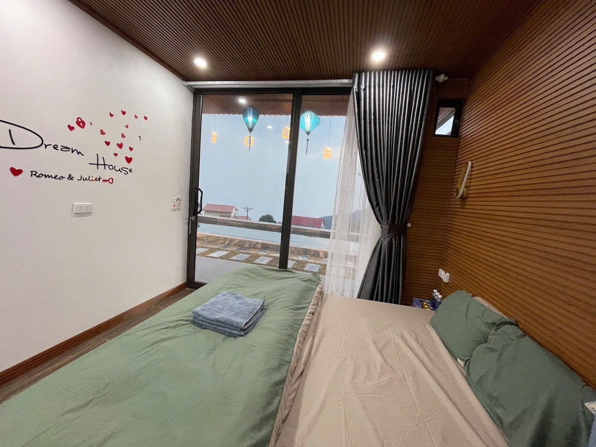 A serene bedroom shows two spacious beds covered in soft linens, with a large glass door that opens to a patio. A decorative wall displays a welcoming message, and light filters through sheer curtains, enhancing the calming ambiance of the space.