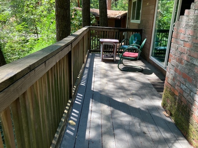 A wooden deck features comfortable seating, including a cushioned chair and a small table, surrounded by trees that provide ample shade. Sunlight filters through the leaves, creating patterns on the deck's surface.