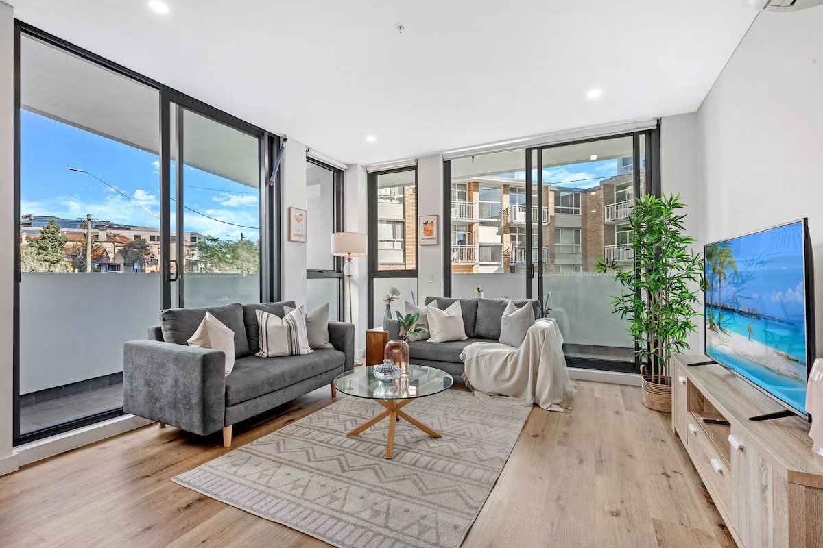 A modern living area features two grey sofas arranged around a glass coffee table. A large television is positioned on a light wood console, while large windows provide ample natural light and a view of the outdoors. Decorative plants are placed strategically in the space.