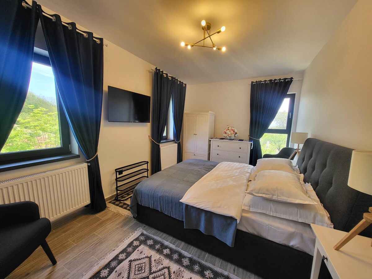 A serene bedroom is highlighted by a spacious bed with layered white and blue bedding. The room features dark curtains framing large windows, and a mounted television is positioned on the wall. A modern lamp and side table accompany a comfortable chair in the corner.