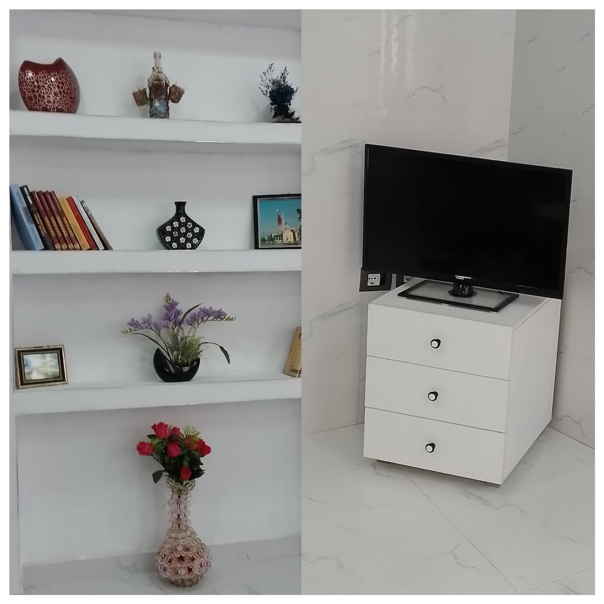 A sleek, modern living area features white floating shelves stocked with books and decorative items. A vibrant floral arrangement is positioned on the bottom shelf. A contemporary TV stand with three drawers sits against a shiny marble wall, providing a functional and aesthetic focal point.