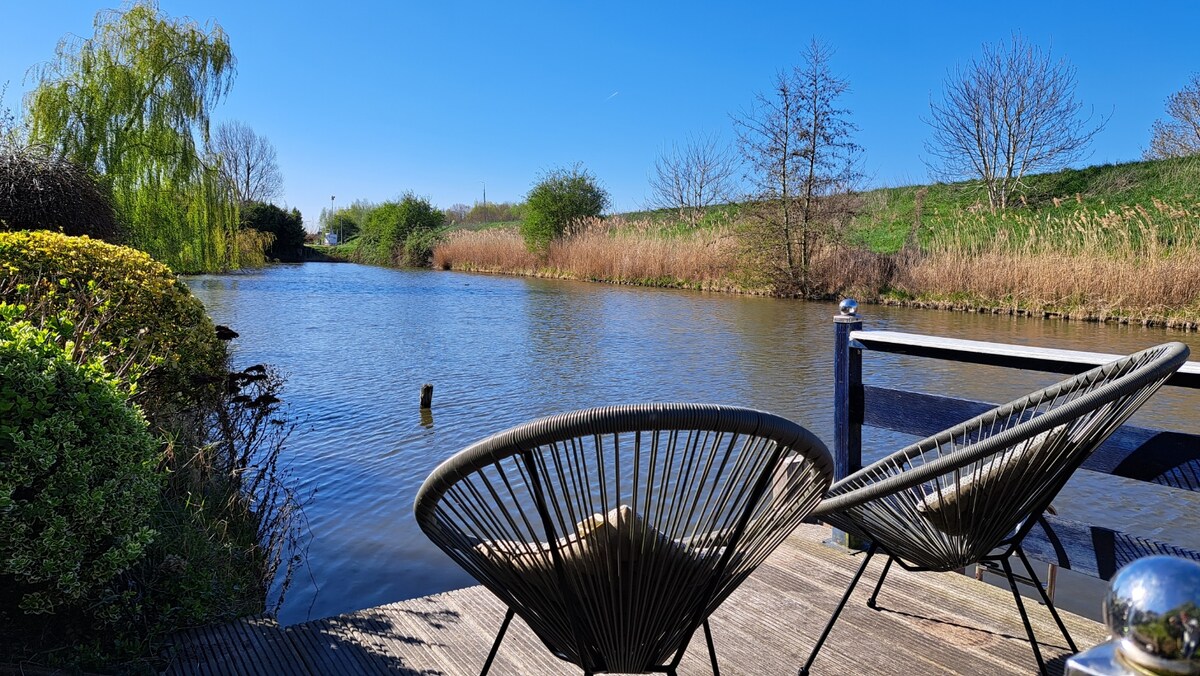 Two modern chairs are positioned near the water's edge, offering views of a serene river surrounded by lush greenery. The calm water surface reflects the clear blue sky, while tall reeds and bushes line the opposite bank, contributing to a tranquil outdoor setting.