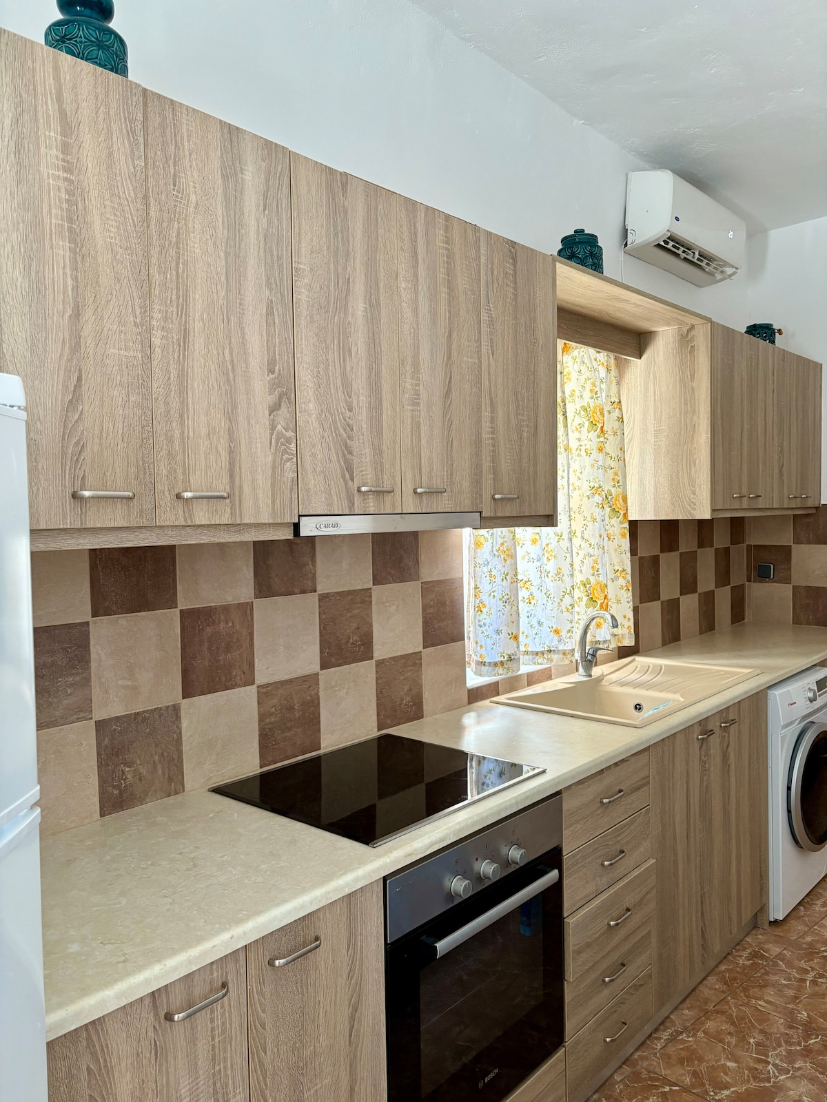 A modern kitchen is showcased, featuring wooden cabinetry and a light stone countertop. A wall-mounted air conditioning unit is visible above the sink, and the floor displays a warm tile pattern. Bright floral curtains allow natural light to illuminate the space.