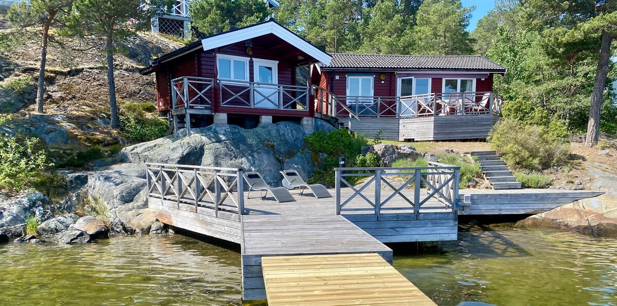 A charming red guest house is situated at the water's edge, surrounded by greenery. A wooden dock extends into the lake, featuring several lounge chairs. Steps lead up from the dock to the elevated building, which includes multiple windows that offer scenic views.
