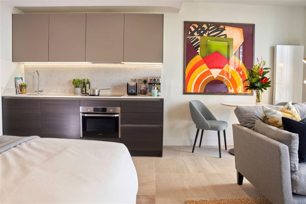 A modern kitchen area is equipped with sleek cabinetry and contemporary appliances. A small dining chair is positioned beside a round table. A vibrant artwork adds a pop of color to the wall. A comfortable sofa, adorned with decorative pillows, complements the inviting space.