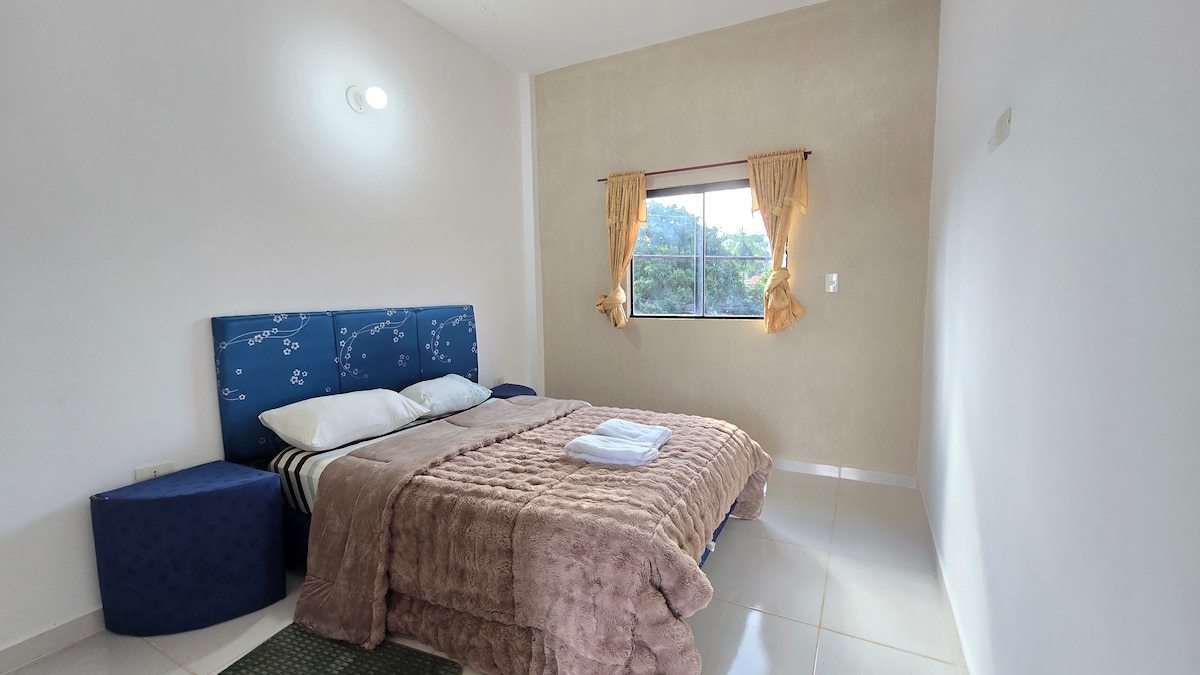 A comfortable sleeping area is highlighted by a plush, textured bedspread over a spacious bed. Two soft pillows rest at the head. Natural light enters through a window dressed with sheer curtains, complemented by a soft-colored wall and cozy seating nearby.