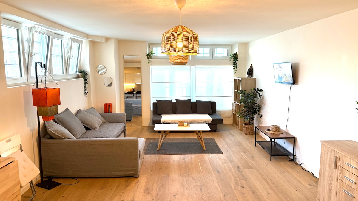 A spacious living area features two comfortable sofas arranged around a central coffee table. Natural light fills the room through large windows, while a television is mounted on the wall. Decorative plants and modern lighting add to the inviting ambiance.
