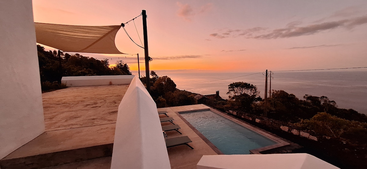 A serene outdoor area features a private pool surrounded by lounge chairs, with gentle waves lapping at the shore in the distance. The setting is enhanced by a beautiful sunset, casting warm hues across the sky, while a canvas shade offers protection from the sun.