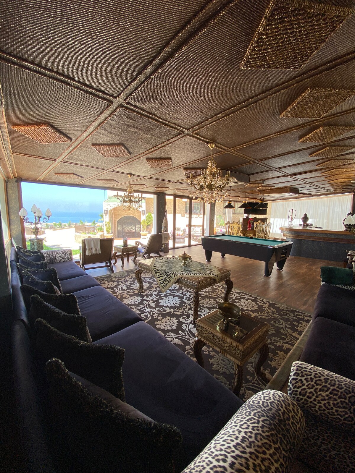 A spacious lounge area features plush seating arrangements with a rich, dark color scheme. Natural light floods the space through large glass doors, revealing views of the sea. A pool table and elegant chandelier add to the ambience, complemented by decorative accents throughout.