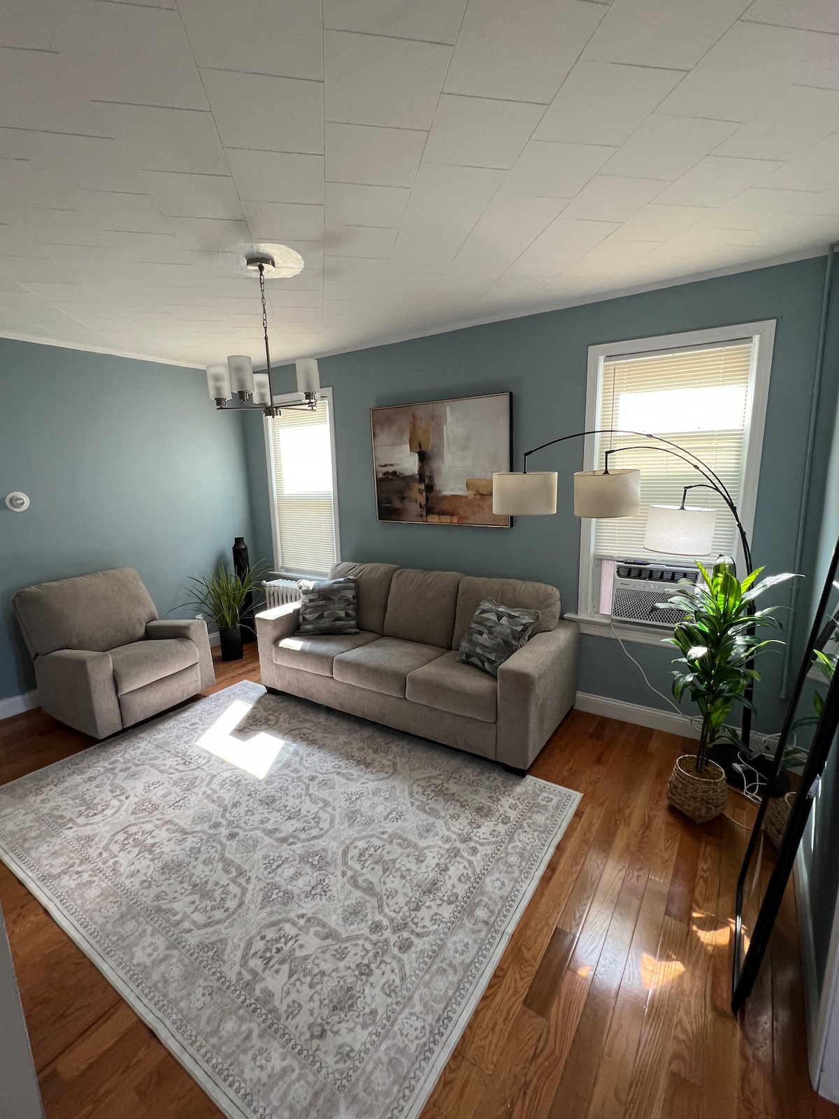 A comfortable living area features a soft beige couch with decorative pillows, accompanied by a reclining chair. Natural light fills the space through a window, where a standing lamp adds illumination. A potted plant sits near the window, enhancing the inviting atmosphere.