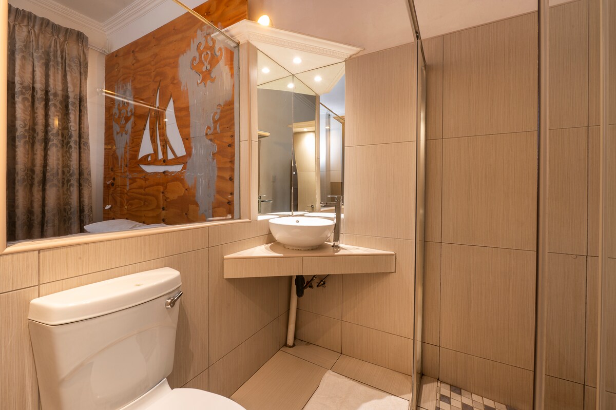A modern bathroom features a sleek toilet and a unique basin sink mounted on a wall. A glass shower enclosure stands beside a mirror, reflecting soft ambient lighting. Neutral-toned tiles cover the floor and walls, creating a clean and functional space.