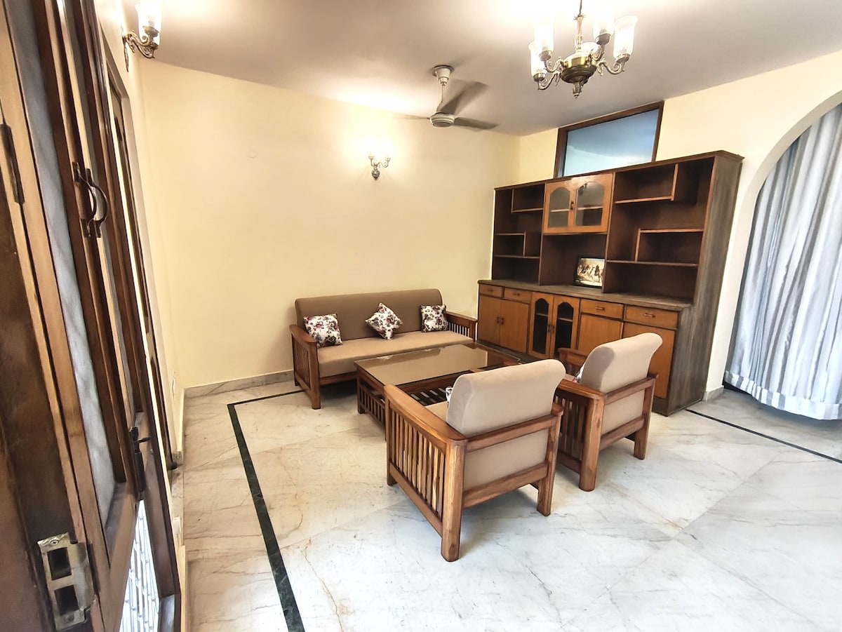 The living area features a wooden seating arrangement with a spacious sofa and two armchairs. A wooden cabinet displays personal items, and large windows allow natural light to fill the space. Cool marble flooring adds a touch of elegance to the room.
