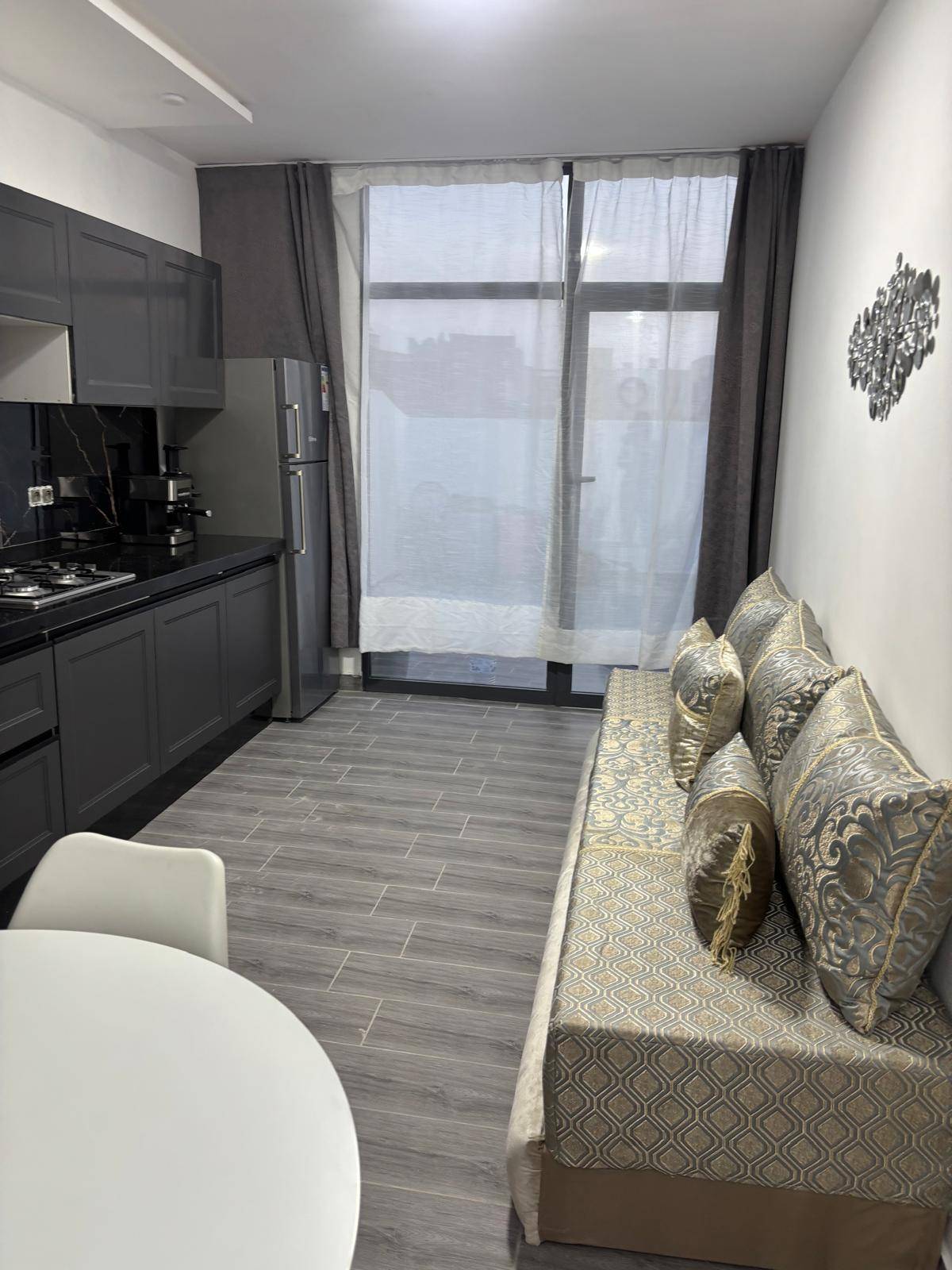 A modern kitchen area is shown with dark cabinetry and a gas stove. A light-colored sofa, adorned with decorative cushions, complements a round dining table. Natural light enters through large windows, enhanced by flowing sheer curtains.