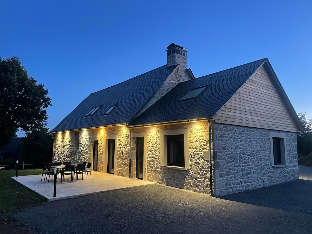 The rustic stone exterior of the house is illuminated by warm lights at dusk, showcasing its modern renovations. Large windows provide glimpses of the interior, while a spacious patio is furnished with a dining table and chairs, set against a tranquil natural backdrop.