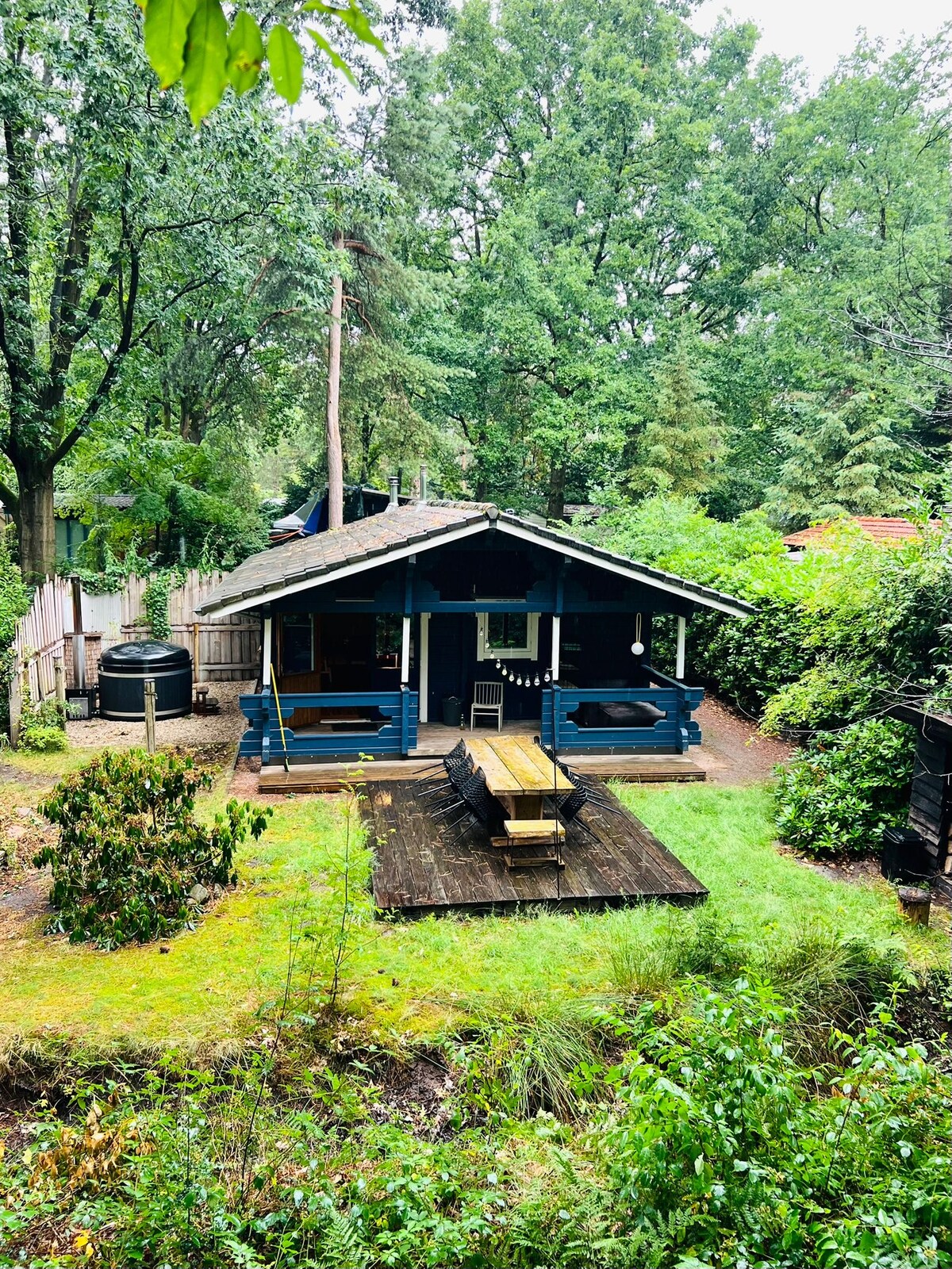 A charming cabin sits nestled among lush green trees, featuring a spacious outdoor dining table on a wooden deck. The structure has a welcoming porch and a hot tub nearby, all surrounded by natural vegetation, creating a peaceful outdoor space.