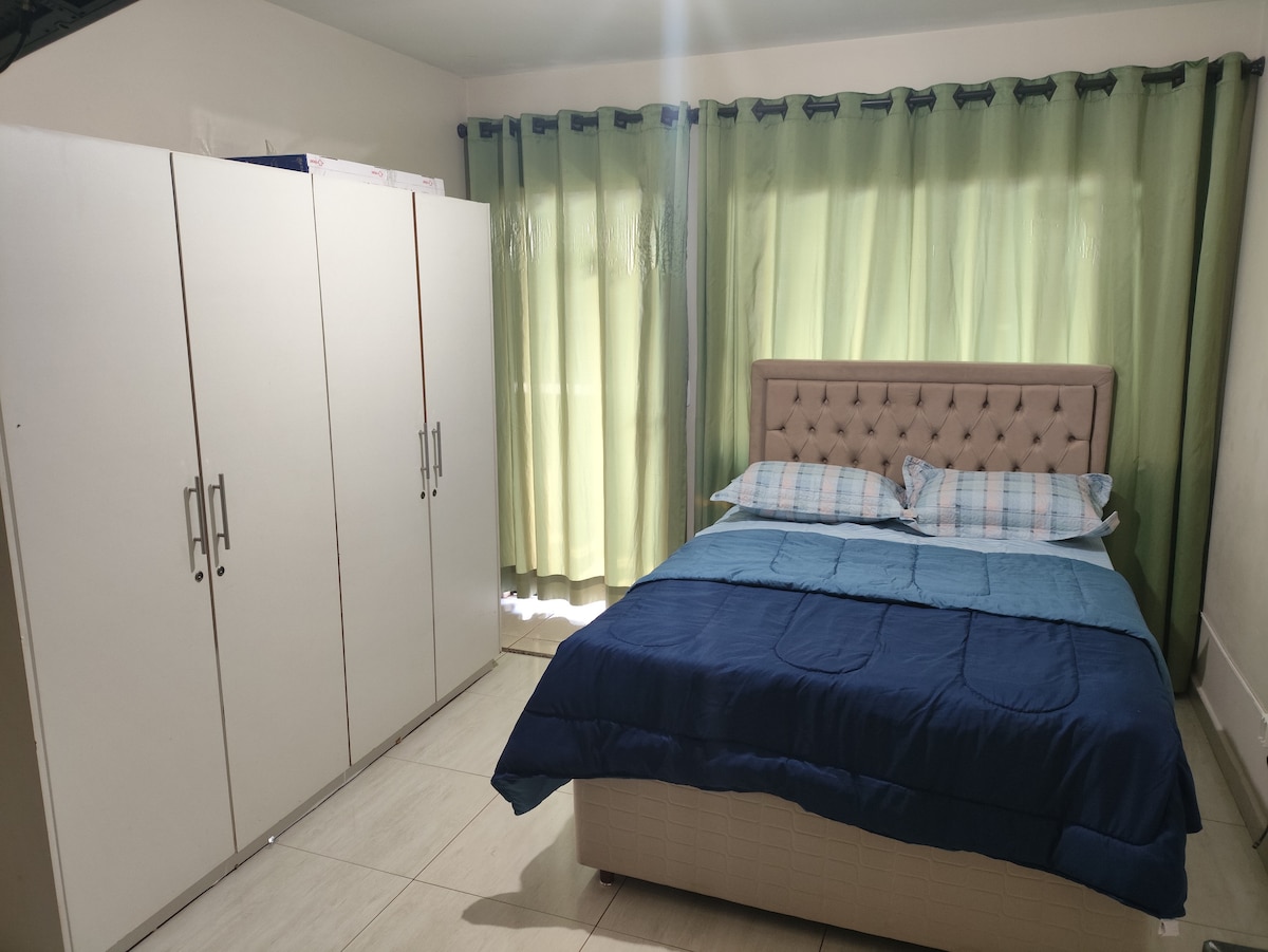 A well-lit sleeping area features a spacious bed with a tufted headboard, dressed in a blue blanket and patterned pillows. A wardrobe with multiple doors stands adjacent to the bed, while light filters through floor-length green curtains.