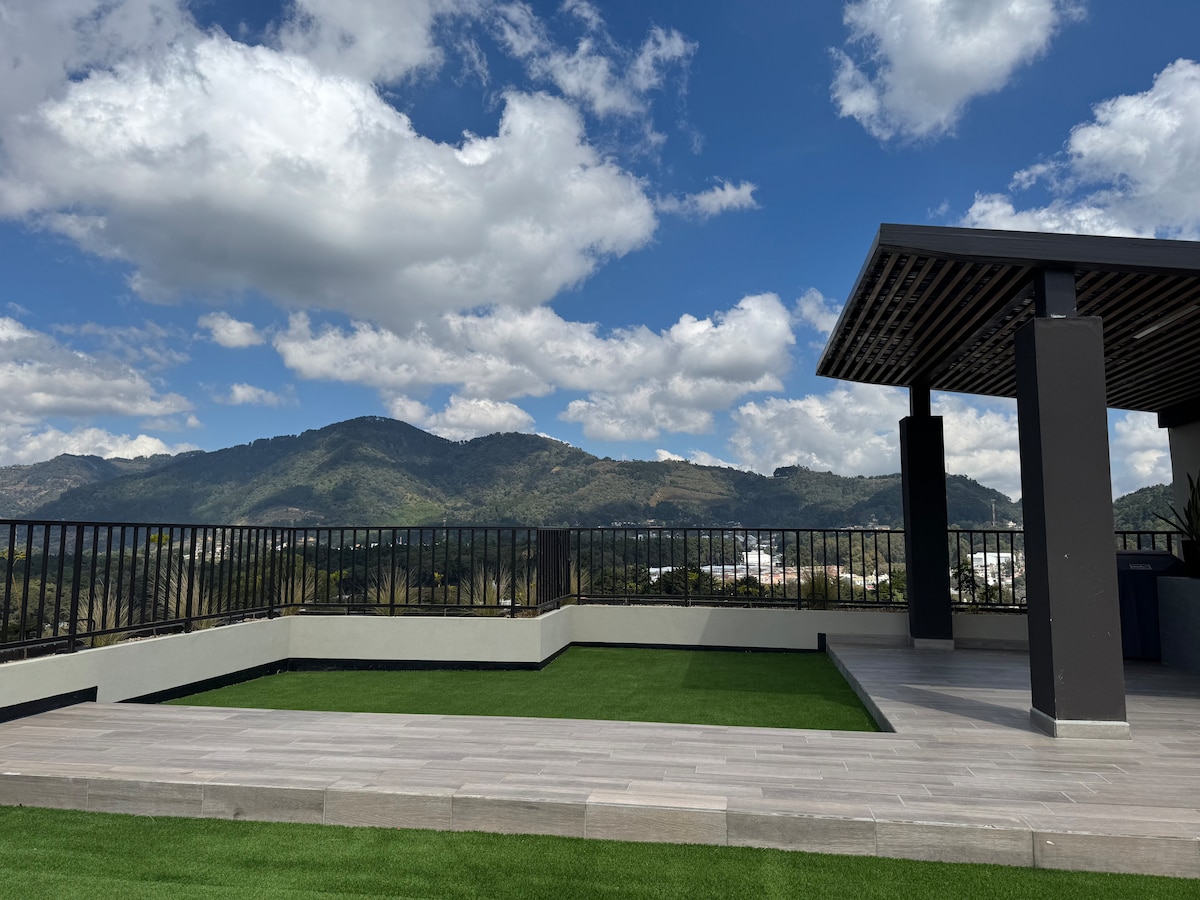 An outdoor terrace features a well-maintained grassy area surrounded by sleek tiles. Mountains can be seen in the background under a partly cloudy sky, enhancing the tranquility of the space. A shaded structure provides a seating area, ideal for relaxation.