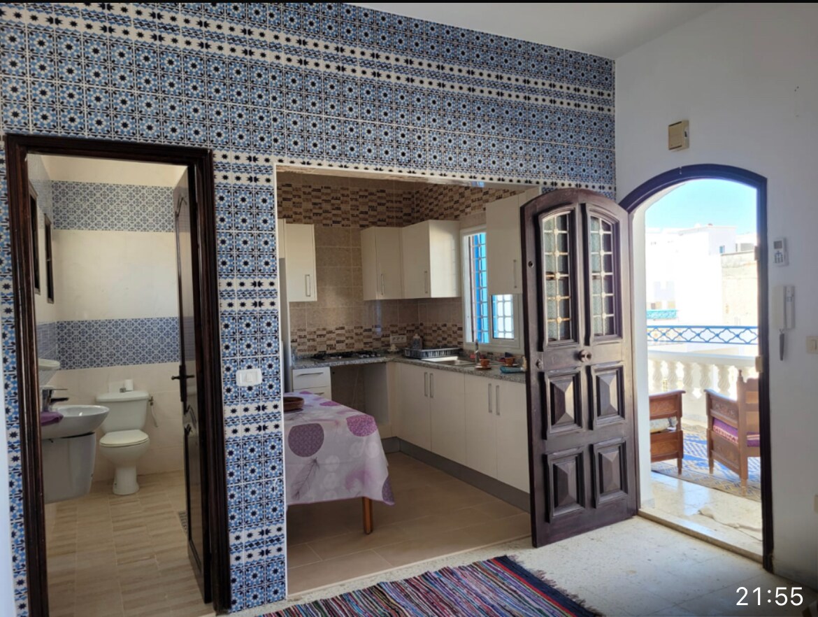 The image presents a bright kitchen area that features patterned tile walls and modern cabinetry. A dining table is set with a tablecloth, while two chairs are positioned nearby. An arched doorway leads to a balcony, which is visible framed by decorative wooden doors.