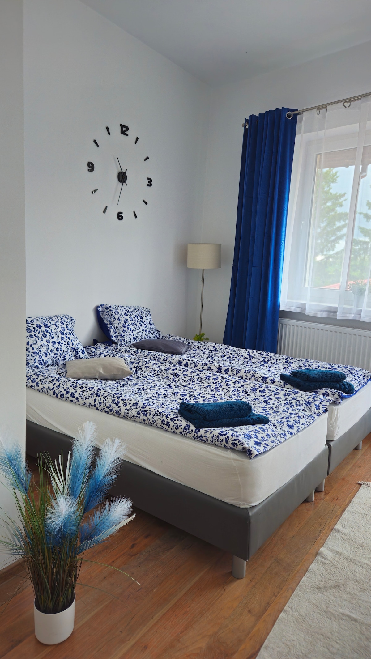 A tranquil sleeping area features a large bed adorned with patterned bedding in soothing blue tones. Two pillows are positioned on each side, while a soft throw is neatly placed at the foot. Natural light filters through the window, complemented by blue drapes and a decorative plant nearby.
