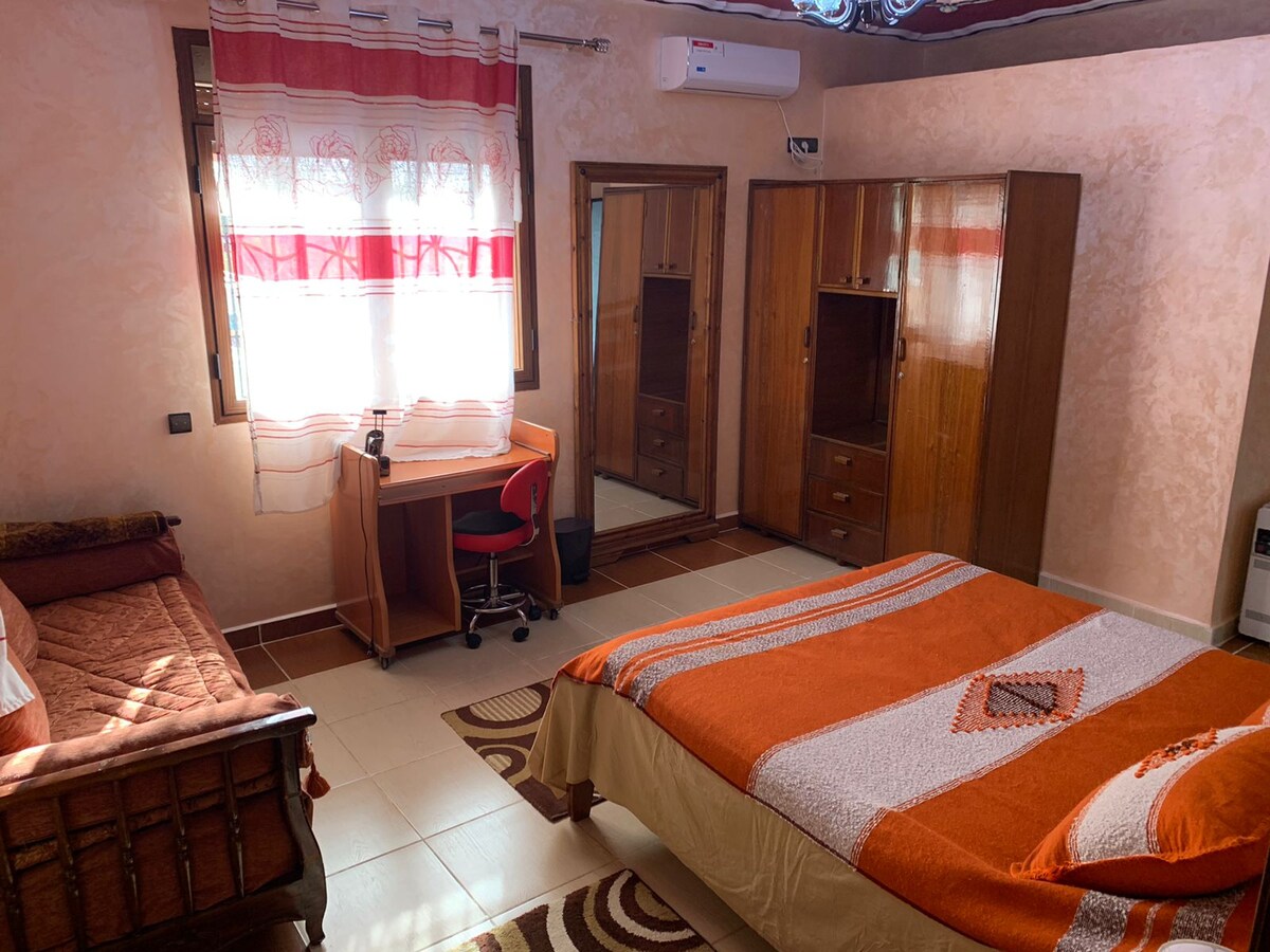 A cozy bedroom is displayed, featuring a spacious bed with an orange and white patterned blanket. A wooden desk with a red chair is positioned beside a large mirror and two wooden wardrobes. Natural light enters through a window adorned with flowing red and white curtains.