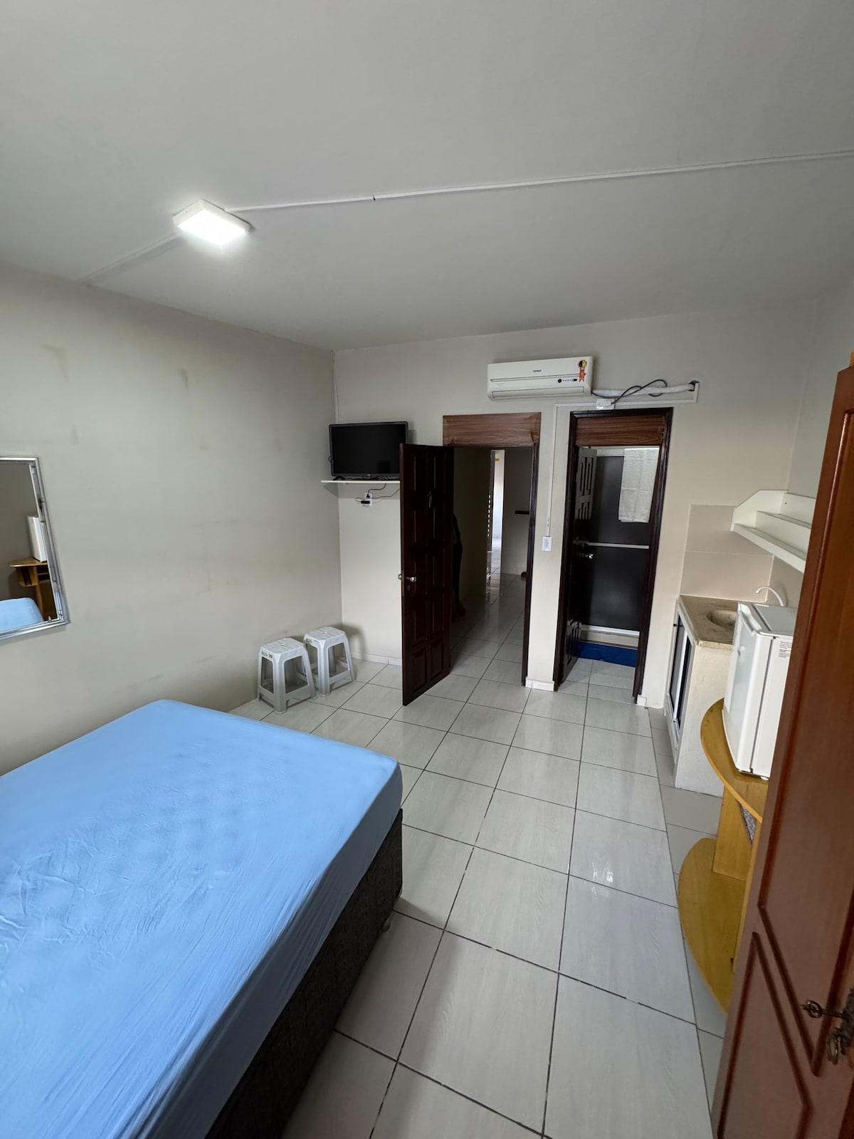 A clean and quiet room features a blue mattress on a dark base and is equipped with a small kitchenette. A mirror is mounted on the wall opposite the bed, and a television is positioned on a shelf nearby. Light from a ceiling fixture enhances the space.