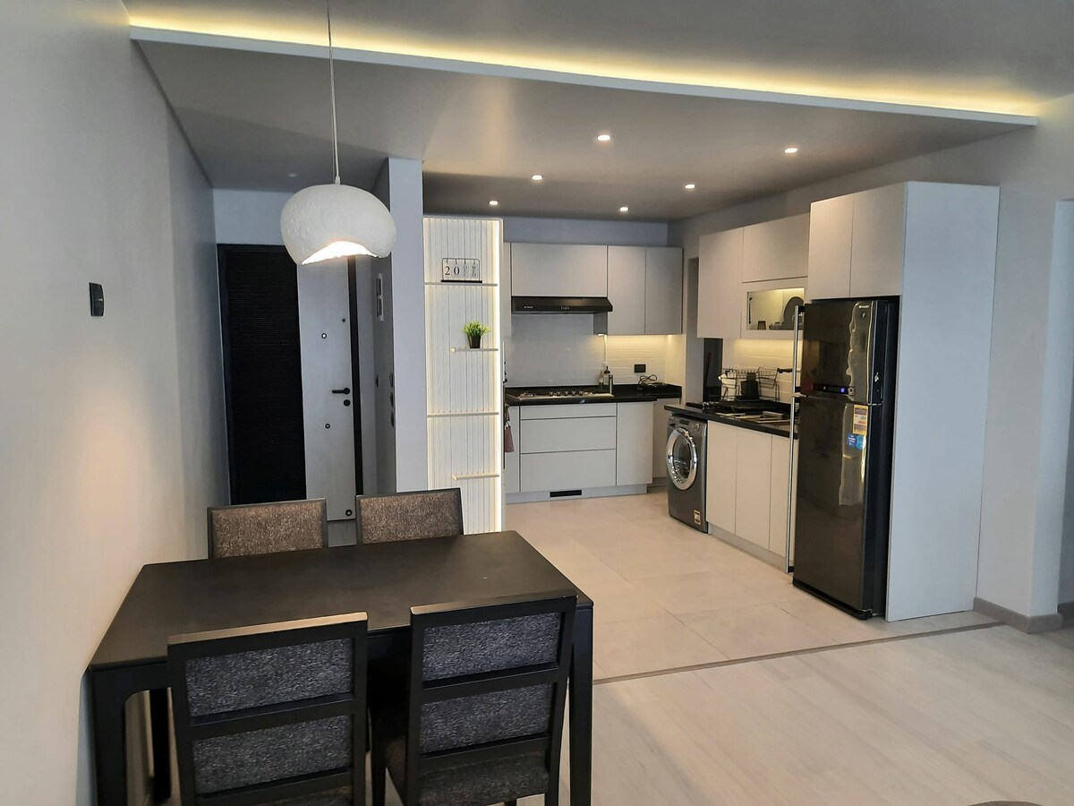 A modern kitchen and dining area is presented with sleek cabinetry and stainless steel appliances. A dining table with four chairs occupies the space. Soft lighting illuminates the room, highlighting the minimalist design and functionality of the layout.