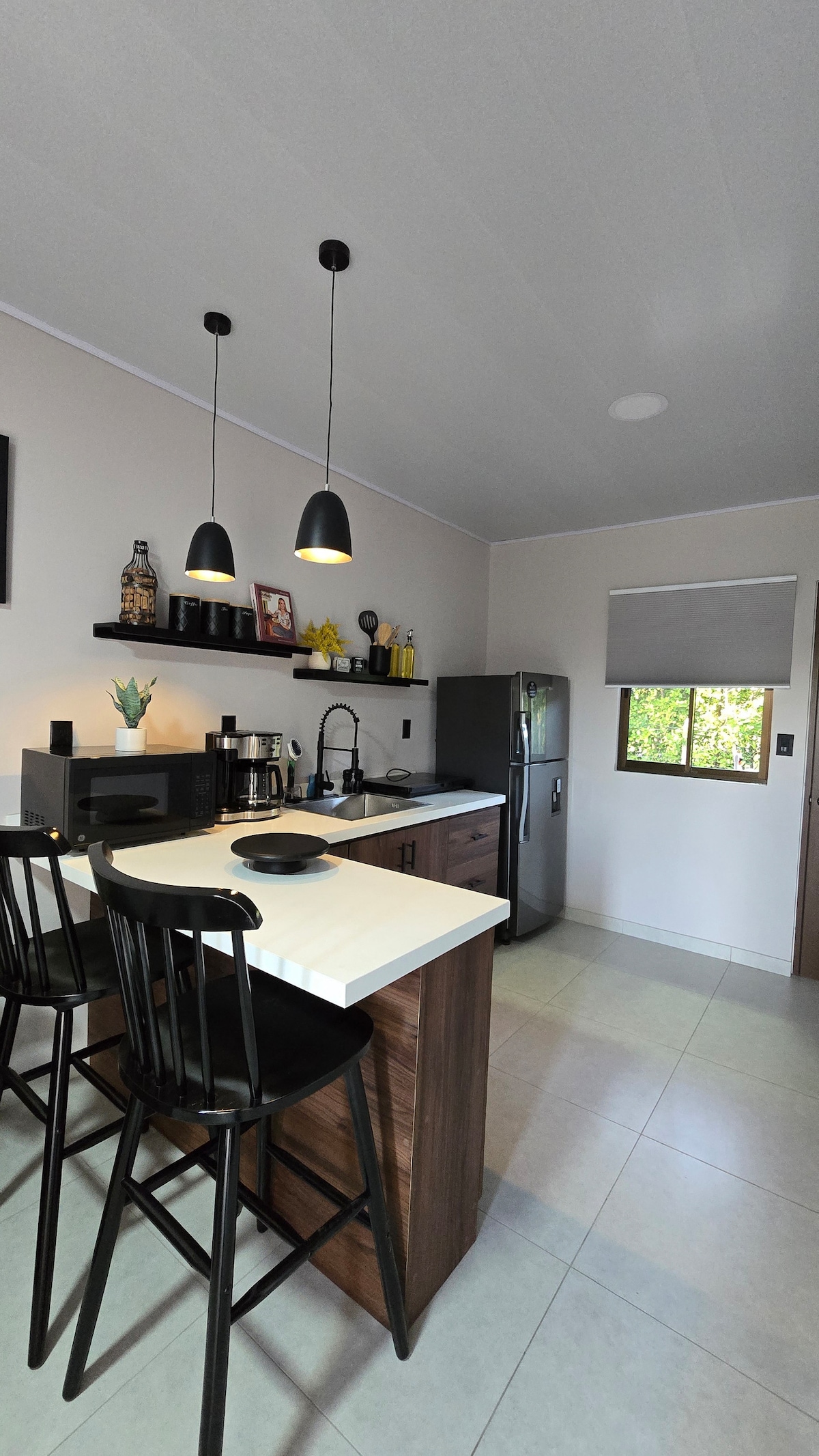 The kitchen area features modern black bar stools positioned at a stylish counter. Sleek appliances, including a refrigerator and coffee maker, are visible, while open shelves hold decorative items and kitchenware. Natural light enters through a nearby window, complemented by light-colored cabinetry.