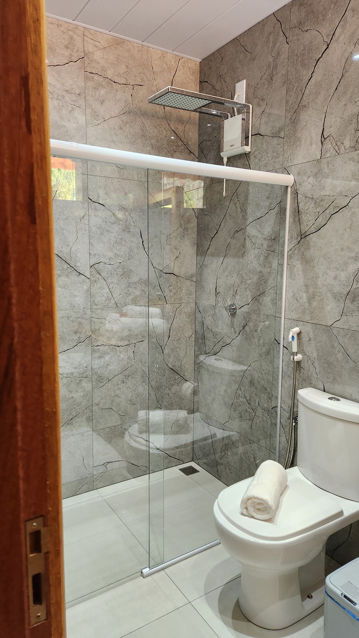 A modern bathroom features a glass shower enclosure with a rainfall showerhead. Grey marble-patterned walls provide a sleek backdrop, while a white toilet and a neatly rolled towel on the sink add functional elements. Natural light filters through a small window.