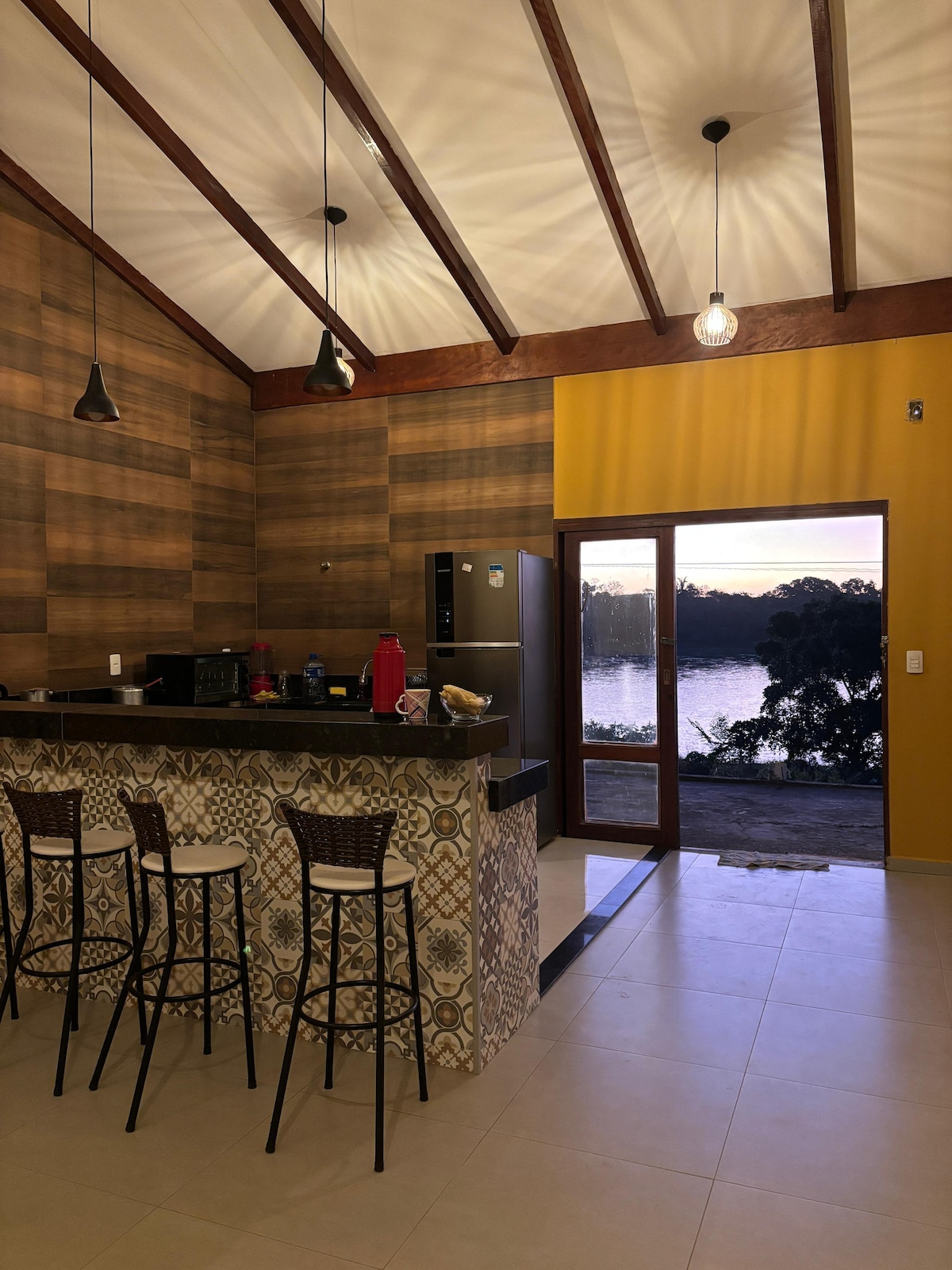 A modern kitchen features a dark countertop with several stools arranged around it. The wooden walls and high ceiling create an open feel. Large glass doors provide a view of the water outside, framed by yellow walls that contrast with the natural wood tones.