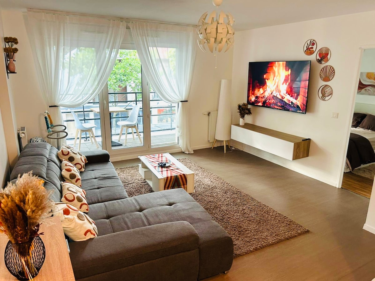 A spacious living area features a modern sectional sofa adorned with patterned cushions. A decorative coffee table occupies the center, while a large TV mounted on the wall displays a simulated fire. Soft light filters through sheer curtains from the balcony access.
