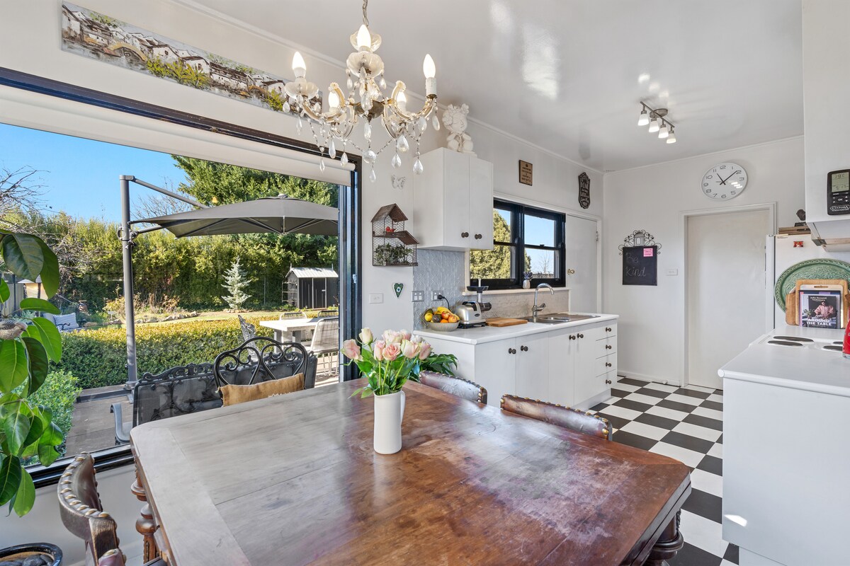 A well-lit kitchen is displayed with black and white checkered flooring. A wooden dining table, adorned with a vase of tulips, sits at the center. Natural light enters through large windows, revealing a garden view and outdoor seating area. Elegant light fixtures are positioned above.