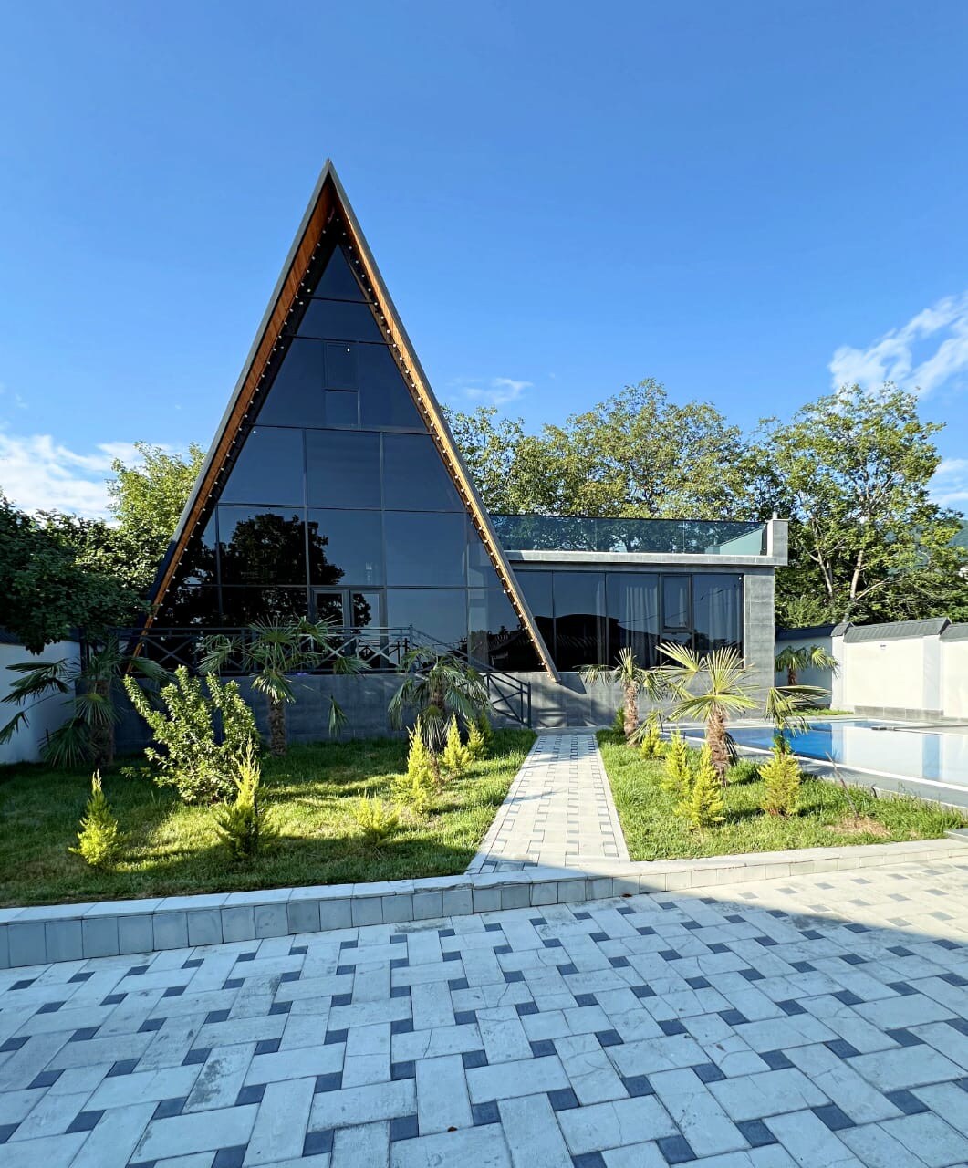 A distinctive triangular structure features expansive glass walls that reflect the sky and surrounding greenery. A landscaped area with low-growing plants borders the building, while a paved walkway leads towards a pool area, creating a harmonious outdoor space.